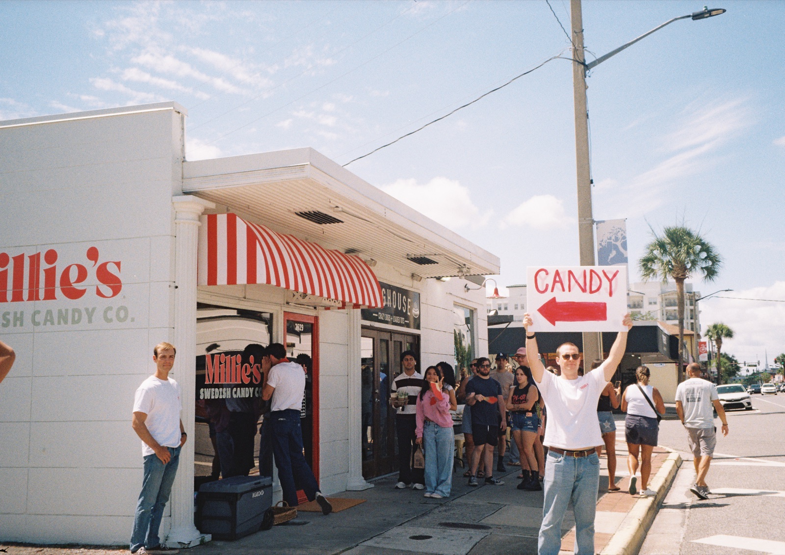 Millie's Swedish Candy Co. on Edgewater Drive: the striped awning, a line of customers out front, and someone holding a hand-painted CANDY sign pointing to the shop.
